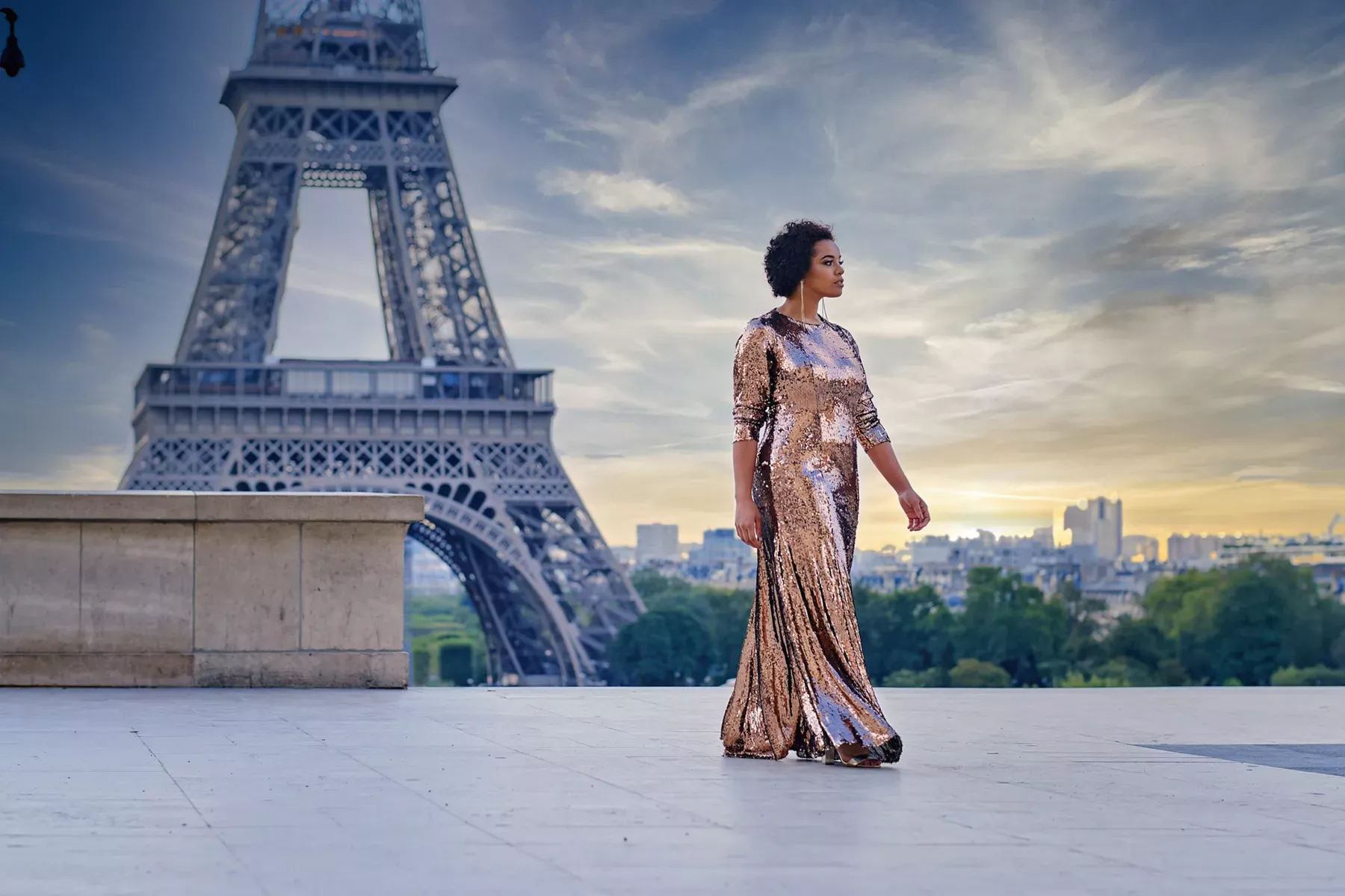 Femme élégante en robe longue à paillettes dorées marchant devant la Tour Eiffel à Paris au coucher du soleil. Scène romantique avec un ciel nuageux et une vue panoramique sur la ville. Photographie de mode et de voyage capturant l'essence de la capitale française.