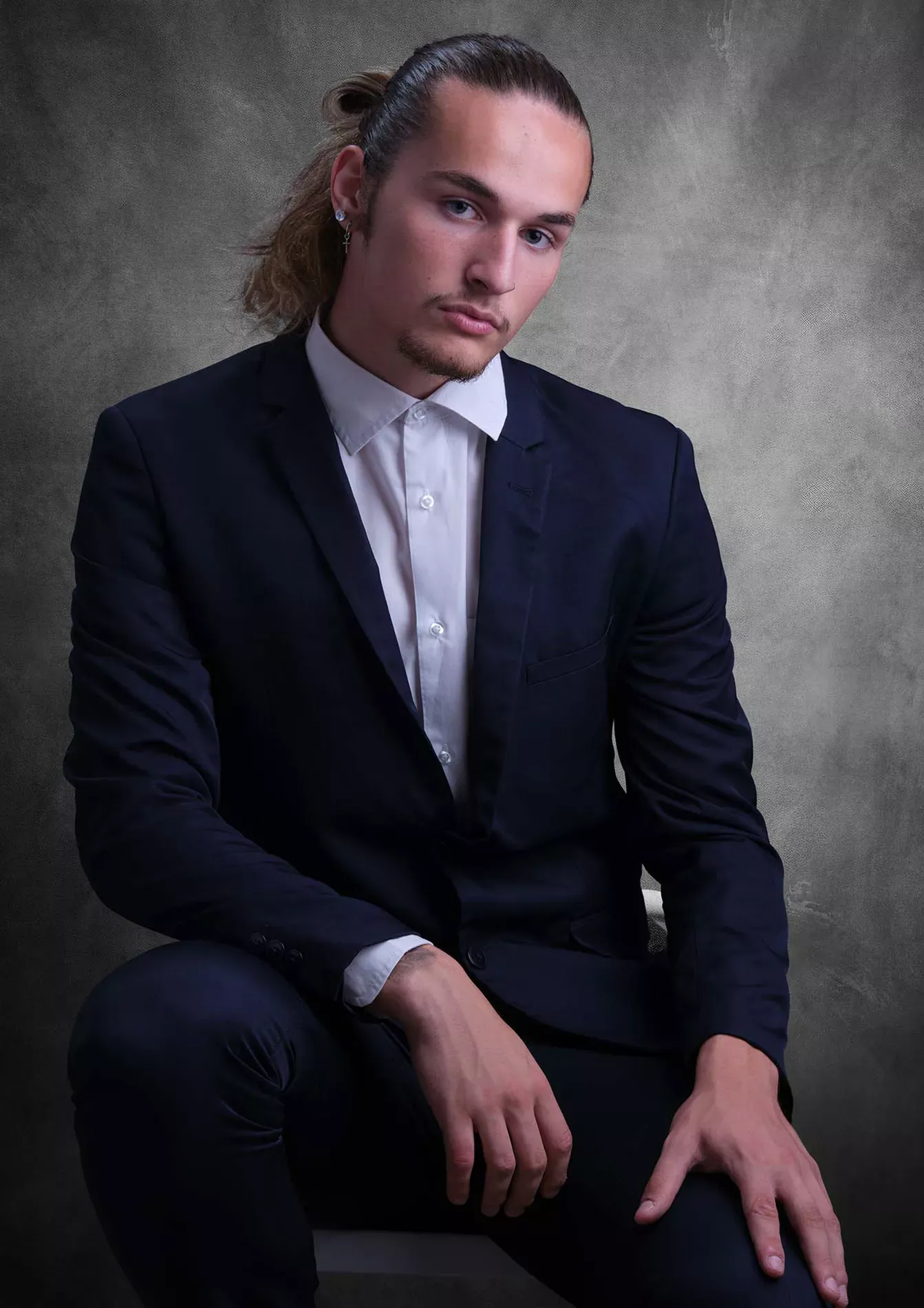 Homme élégant en costume bleu marine et chemise blanche, assis en studio avec une coiffure en chignon, photographié par TRIFA Studio. Portrait professionnel sur fond gris texturé, mettant en valeur un style moderne et sophistiqué