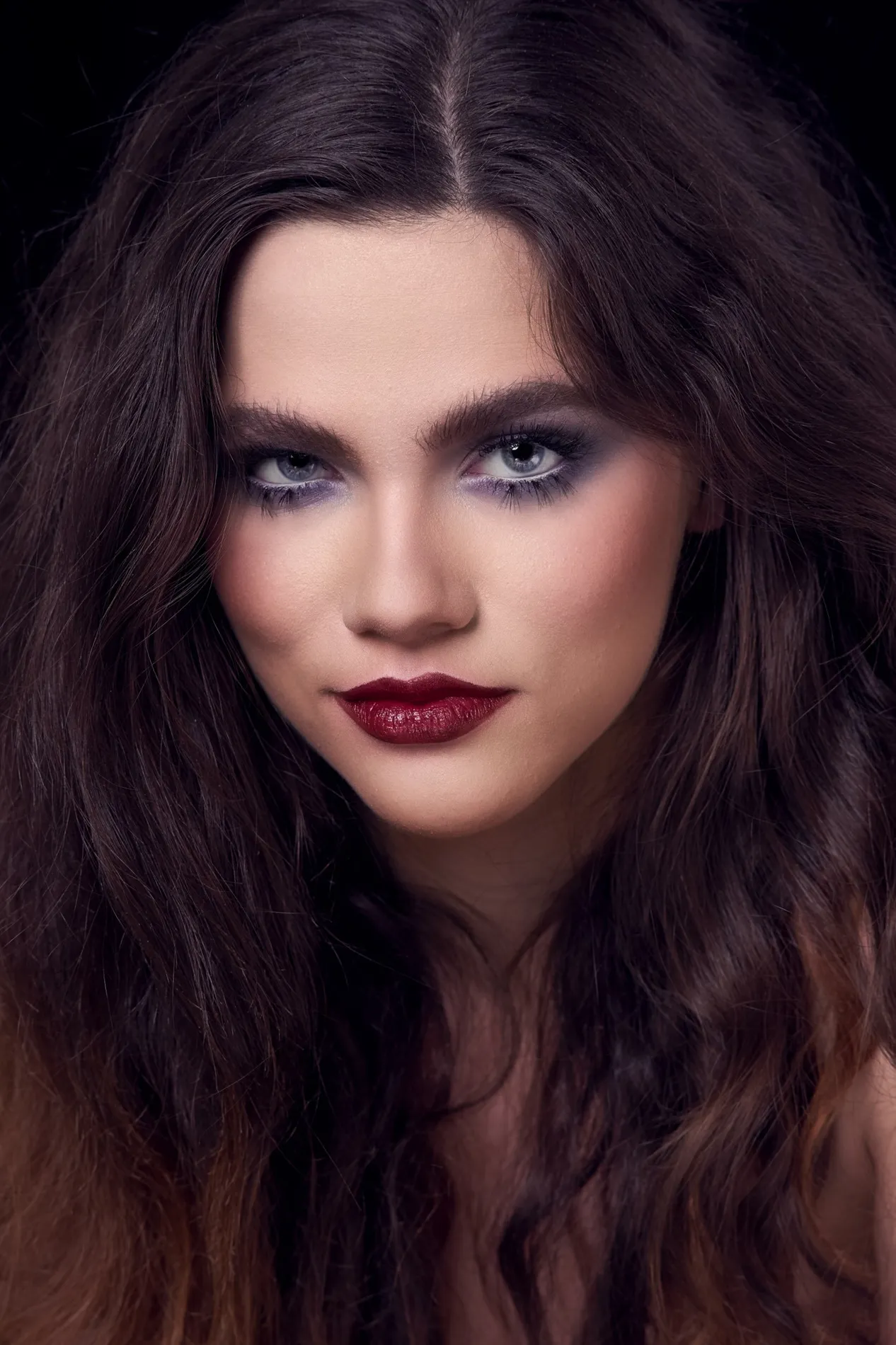 Portrait glamour d’une femme aux cheveux bruns ondulés sur fond noir, capturé par TRIFA studio. Mise en beauté sophistiquée avec un maquillage audacieux, mettant en valeur des lèvres rouge foncé et des yeux soulignés de fard violet. Photographie professionnelle de mode et de portrait, idéale pour découvrir le talent de TRIFA Studio sur son site officiel.