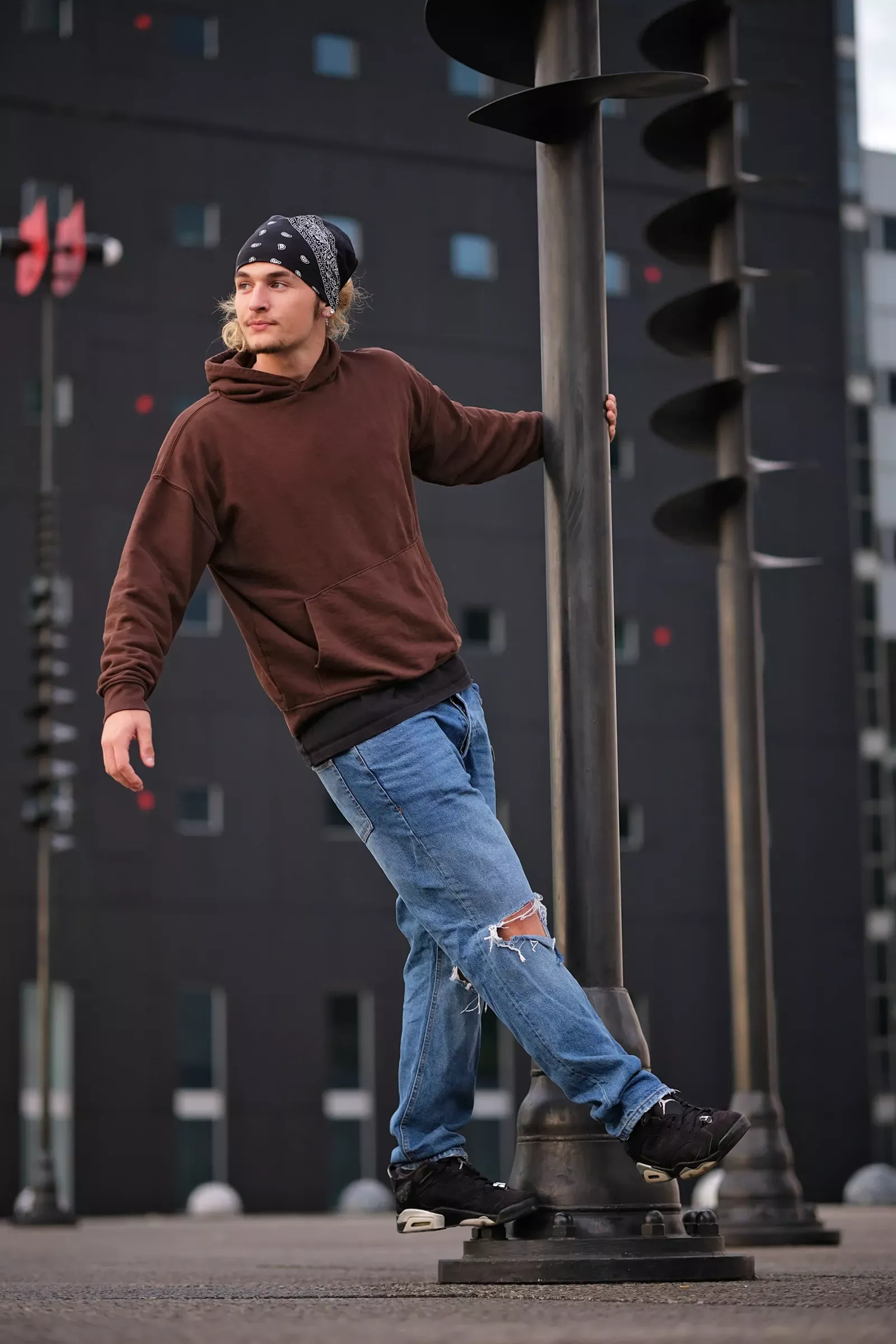 Portrait extérieur captivant d’un jeune homme stylé en tenue urbaine, posant avec assurance contre un poteau métallique décoratif. Vêtu d’un hoodie marron élégant, d’un bandana noir à motifs blancs et d’un jean bleu déchiré, il incarne un look moderne. Arrière-plan contemporain avec un bâtiment aux façades sombres et fenêtres rectangulaires. Photo réalisée par TRIFA Studio, expert en portraits extérieurs.