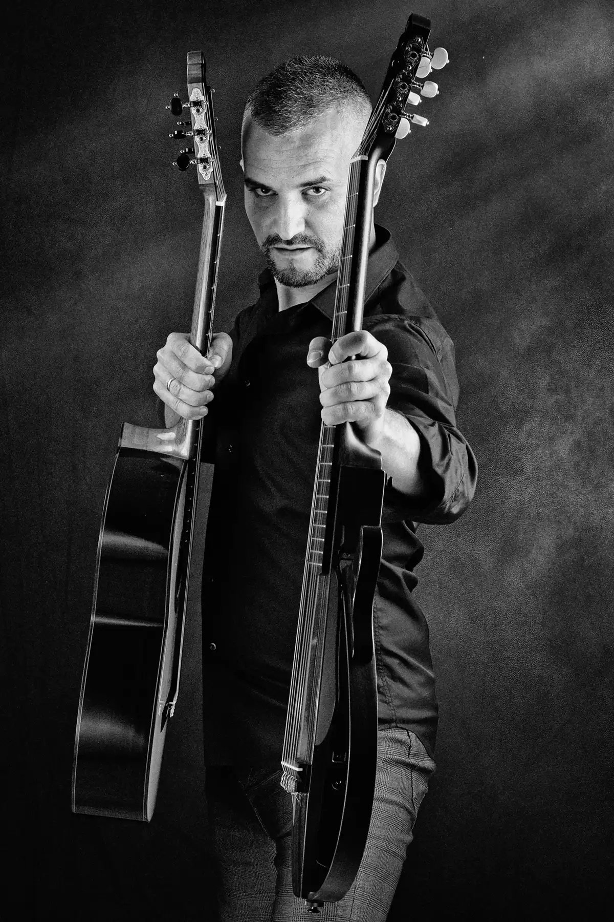 Portrait en noir et blanc d’un musicien tenant deux guitares en studio, photographié par TRIFA Studio. Homme en chemise noire et pantalon à carreaux, capturé dans une pose dynamique sur fond sombre. Photographie artistique mettant en valeur la passion pour la musique.
