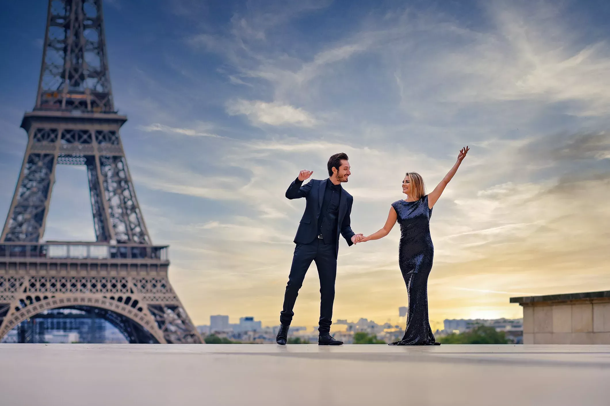 Couple élégant dansant près de la Tour Eiffel au coucher du soleil à Paris. Homme en costume noir et femme en robe à sequins noirs se tenant la main et célébrant avec joie. Photographie romantique de voyage et de mode en France.