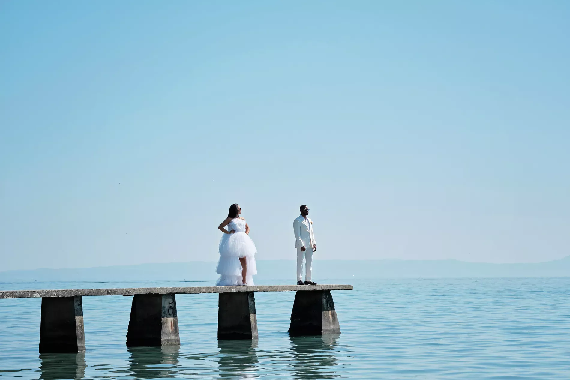 Magnifique séance photo de mariage en extérieur réalisée par TRIFA Studio au bord du Lac Léman. Un couple de mariés pose sur un ponton en béton, entouré d'eau calme et d'un ciel bleu éclatant. La mariée porte une élégante robe blanche volumineuse, tandis que le marié arbore un costume clair raffiné. L'ambiance est minimaliste et romantique, mettant en valeur la beauté naturelle du paysage et l’amour du couple. Photographie artistique et professionnelle, parfaite pour immortaliser un moment unique.
